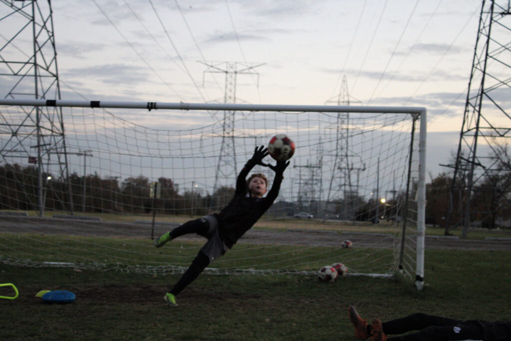 Goalkeeper reaching upward in a high dive to block a powerful top-corner shot